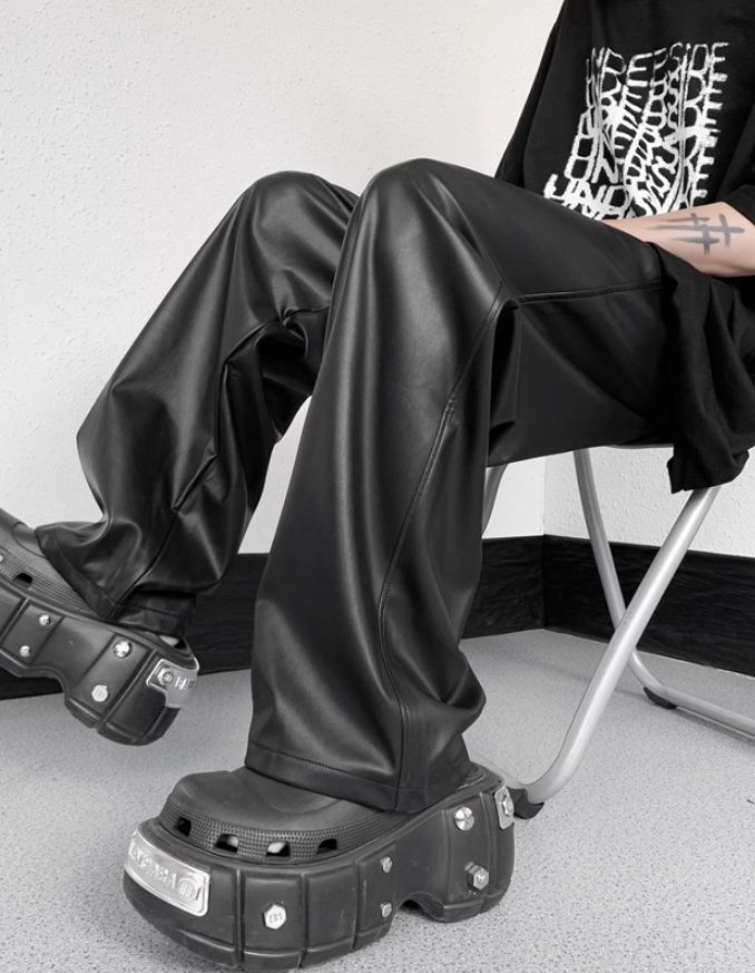 Person sitting on a wooden stool wearing loose-fitting black faux leather pants, paired with a white shirt and black jacket in a mirror selfie setup.