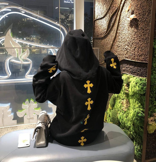 Black hoodie with yellow cross graphics visible on the center back and sleeves, worn by a person sitting indoors.