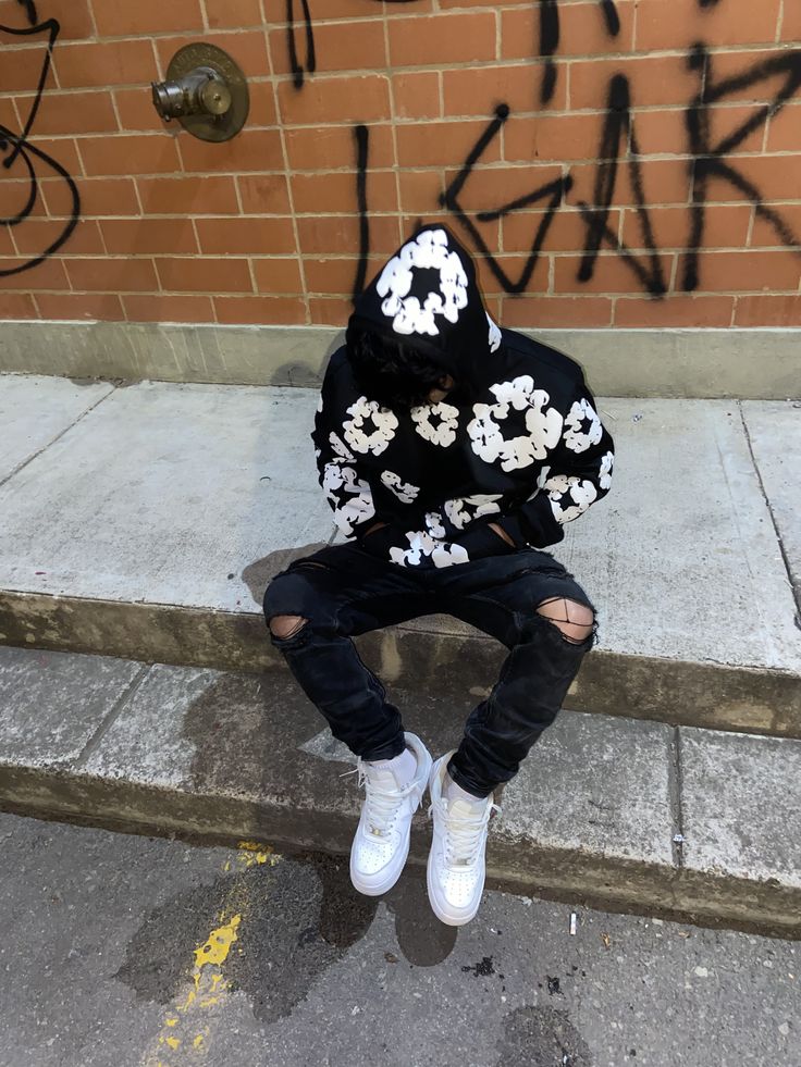 Person wearing a black and white floral hoodie sitting on a sidewalk with graffiti in the background
