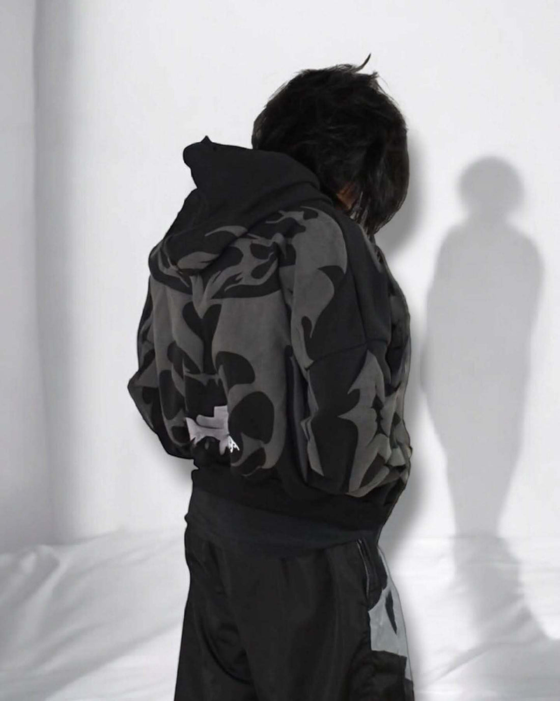 Person wearing a black and gray hoodie with a logo, standing against a white wall.