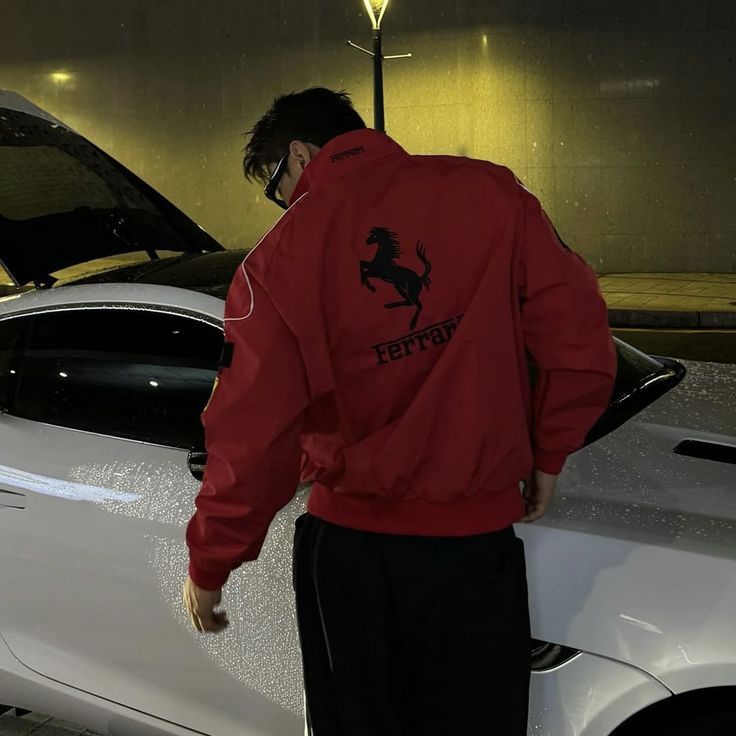 Person wearing a red Ferrari jacket standing next to a car at night.